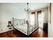 Schlafzimmer mit einem schwarzen Metalldoppelbett mit geschwungenem Design, Holzboden, weißen Wänden und zwei großen Fenstern mit weißen Vorhängen und Holzjalousien. Neben dem Bett stehen ein runder Beistelltisch und ein kleiner Schrank.