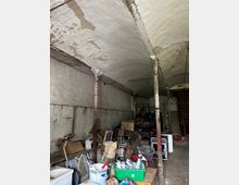 Ein verlassener Lagerraum mit gewölbter Decke und metallenen Stützpfeilern, die deutliche Rostspuren aufweisen. Der Raum ist mit Gegenständen, Kartons und Werkzeugen unordentlich gefüllt, die Wände zeigen Abnutzung und abblätternde Farbe.