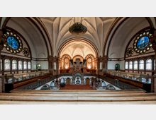 Innenansicht einer Kirche mit einer hohen, gewölbten Decke und detaillierten Backsteinstrukturen. Über dem Altarbereich befindet sich eine große Orgel, flankiert von verzierten Fenstern und Statuen; bunte Rosettenfenster und Reihen von Holzbänken prägen den Raum.