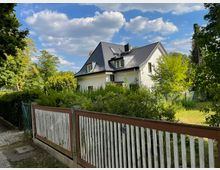 Haus mit weißer Fassade und dunkelgrauem Satteldach inmitten eines Gartens mit Bäumen und Sträuchern. Im Vordergrund ein Zaun aus Holzlatten und ein gepflasterter Weg.