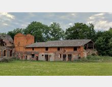 Altes Backsteingebäude mit verwittertem Dach und teilweise eingestürzten Abschnitten, umgeben von Wiese und Bäumen; links erkennt man eine Ruine aus Stein.