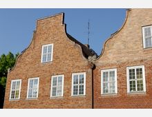 Backsteinfassade eines Gebäudes mit dekorativen geschwungenen Giebeln im Stil der norddeutschen Architektur. Die Fassade hat weiße Fensterrahmen und ist vor einem klaren blauen Himmel abgebildet, mit einem Baum auf der linken Seite.