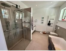 Modernes Badezimmer mit einer großen, verglasten Doppeldusche, einer Badewanne, einem Wand-WC und einem weißen Waschtisch. Die Wände sind weiß, Böden und Duschwände sind mit großen, grauen Fliesen verkleidet; kleine Fenster mit farbigem Glas lassen Tageslicht herein.