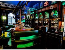 Innenraum einer Bar mit gebogener Holztresenanlage, grünen Polsterbänken und Barhockern sowie hängenden Industrielampen und Metallträgern an der Decke. An der Rückwand stehen beleuchtete Regale mit Flaschen und Gläsern in bogenförmigen Nischen, umgeben von Schildern und Neonwerbung.