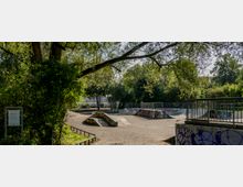 Eine Skateboardanlage im Freien mit Rampen und Halfpipes, umgeben von Bäumen und dichtem Grün. Im Vordergrund sind Geländer und Graffiti sichtbar, die Anlage liegt in einer sonnigen Umgebung.