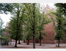 Backsteingebäude mit Spitzgiebeln im Hintergrund, umgeben von großen Laubbäumen. Im Vordergrund ist ein gepflasterter Hof mit Sitzbänken und Tischen zu sehen.