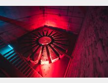 Industrieähnlicher Raum mit einem großen, runden, radial angeordneten Steinelement in der Mitte, beleuchtet in roten und blauen Farbtönen. Eine Metalltreppe führt hinunter, und an der Backsteinwand ist eine Leiter zu sehen.