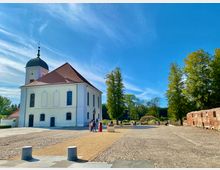 Kirche mit weißer Fassade und rotem Dach, flankiert von einem Glockenturm mit schwarzer Kuppel, umgeben von einem gepflasterten Hof und grünen Bäumen; rechts im Bild eine rote Backsteinmauer. Vor der Kirche sind einige Menschen zu sehen, die das sonnige Wetter genießen.