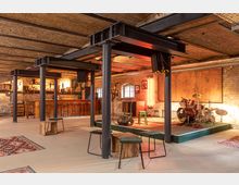 Ein industriell gestalteter Veranstaltungsraum mit Backsteinmauern, freiliegenden Deckenbalken und schwarzen Stahlstützen. Im Vordergrund befinden sich Hocker und Holztische, im Hintergrund eine Bühne mit Schlagzeug sowie eine Bar mit Hockern und Beleuchtung.