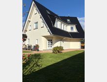 Ein modernes Einfamilienhaus mit heller Fassade und dunkelgrauem Satteldach, großen Fenstern und einem gepflegten Garten mit Rasenfläche im Vordergrund. Im Eingangsbereich befinden sich eine weiße Tür und ein gepflasterter Weg, umgeben von Blumen und Sträuchern.