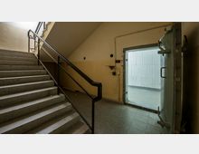 Treppenhaus mit einer Beton-Metalltreppe, Geländer aus schwarzem Metall und einer seitlichen Wand in beigefarbener Farbe. Rechts ist eine massive, geöffnete Stahltür sichtbar, die in einen Raum mit Betonwänden führt.