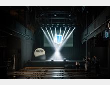 Industriell gestalteter Veranstaltungsraum mit dunklen Wänden, einer Bühne im Hintergrund und leeren Holzbänken im Vordergrund. Über der Bühne sind Scheinwerfer und eine helle Projektion auf einer Leinwand zu sehen, flankiert von Personen am rechten Rand des Bildes.