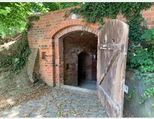 Backsteinbau mit Eingang zu einem Gewölbekeller, umgeben von Efeu und Pflanzen. Eine hölzerne Tür steht halb geöffnet neben dem gemauerten Torbogen, der in den schattigen Innenraum führt.