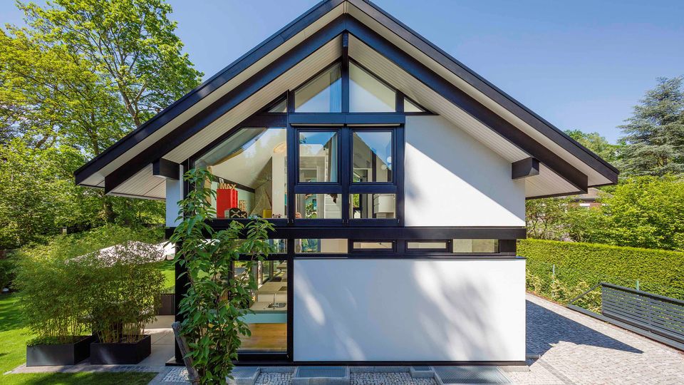 Moderne Wohnhausfassade mit steilem Satteldach, weißer Verkleidung und schwarzen Rahmen sowie großen, dreieckigen Glasflächen im Giebel. Vor dem Haus liegen gepflasterte Wege und Rasenflächen, umgeben von Hecken und hohen Bäumen.