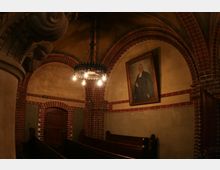 Innenraum einer Kirche mit Gewölbedecke aus Ziegelsteinen, hölzernen Sitzbänken und einem Gemälde eines Geistlichen an der Wand. Im Zentrum hängt ein dekorativer Kronleuchter aus Metall.