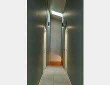 Schmaler, moderner Flur mit graugrünen Wänden, hellen Fliesenboden und zwei offenen Türen auf beiden Seiten. Am Ende des Flurs befindet sich eine Holztreppe, daneben eine Wand aus hellem Holz, die von einem Oberlicht beleuchtet wird.