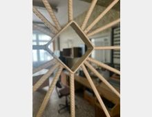 Blick durch ein weißes, gitterartiges Fensterelement mit rautenförmiger Öffnung in einen Büroraum mit Schreibtischen und Stühlen. Im Hintergrund sind große Fenster mit Tageslicht sowie Regale und Schränke zu erkennen.