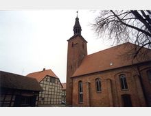 Backstein-Kirche mit spitzem Glockenturm und roten Ziegeldächern, umgeben von Fachwerkhäusern; im Vordergrund ein kahler Baum.