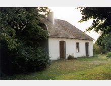 Weißes, einstöckiges Bauernhaus mit Schindeldach, zwei Holztüren und einem kleinen Fenster, umgeben von grünem Rasen und Bäumen. Das Gebäude liegt in einer ländlichen, parkähnlichen Umgebung.