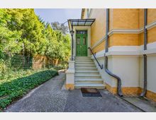 Seitlicher Eingangsbereich eines Backsteingebäudes mit einer Treppe zu einer grünen Doppeltür unter einem Glasvordach. Daneben verläuft ein gepflasterter Weg entlang eines Zauns und dichter Bepflanzung mit Bäumen und Bodendeckern.