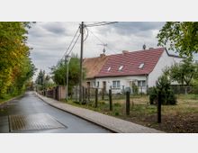 Eine ruhige Wohnstraße mit zwei aneinandergereihten Einfamilienhäusern, die rote Ziegeldächer und helle Fassaden haben. Links der Straße verläuft eine Baumreihe, während rechts Zäune die Vorgärten der Häuser abgrenzen.