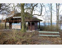 Holzhütte mit Schildern der "Stein-Kreisschifffahrt" in einem parkähnlichen Bereich, umgeben von kahlen Bäumen. Im Vordergrund steht eine grüne Bank, im Hintergrund ist ein Gewässer sichtbar.