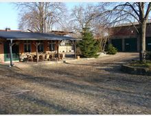 Hof mit Kopfsteinpflaster, einem kleinen Gebäude mit Veranda und Holzstühlen rechts im Bild, sowie mehreren Bäumen und einem Nebengebäude mit grünen Toren im Hintergrund. Die Umgebung wirkt ländlich und offen.