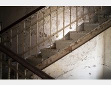 Eine abgenutzte Treppe in einem verfallenen Gebäude mit abblätternder Farbe auf den Stufen, dem Geländer und der Wand. Die Metallstreben des Geländers haben ein gedrehtes Design, und die Umgebung zeigt deutliche Zeichen von Verfall.