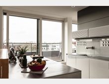 Moderne Küche mit großen, bodentiefen Fenstern, die auf eine Terrasse mit Geländer und Blick auf eine städtische Umgebung führen. Die Einrichtung umfasst eine Kücheninsel mit Edelstahloberfläche, eine rote Obstschale, Kochutensilien und eine minimalistische Schrankwand mit integrierter Dunstabzugshaube.