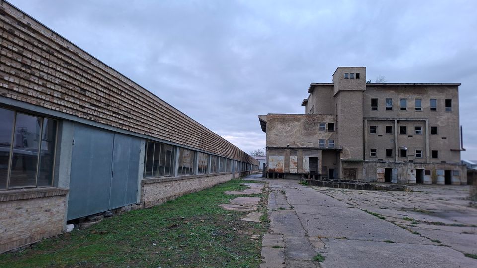Industriegelände mit verwitterten Gebäuden aus Ziegelstein und Beton. Ein langes flaches Gebäude mit großen Fenstern steht links, während rechts ein mehrstöckiges, stark gealtertes Gebäude mit zerbrochenen Fenstern und abgeschlagenen Türen zu sehen ist; der Boden besteht aus Betonplatten mit vereinzeltem Grasbewuchs.