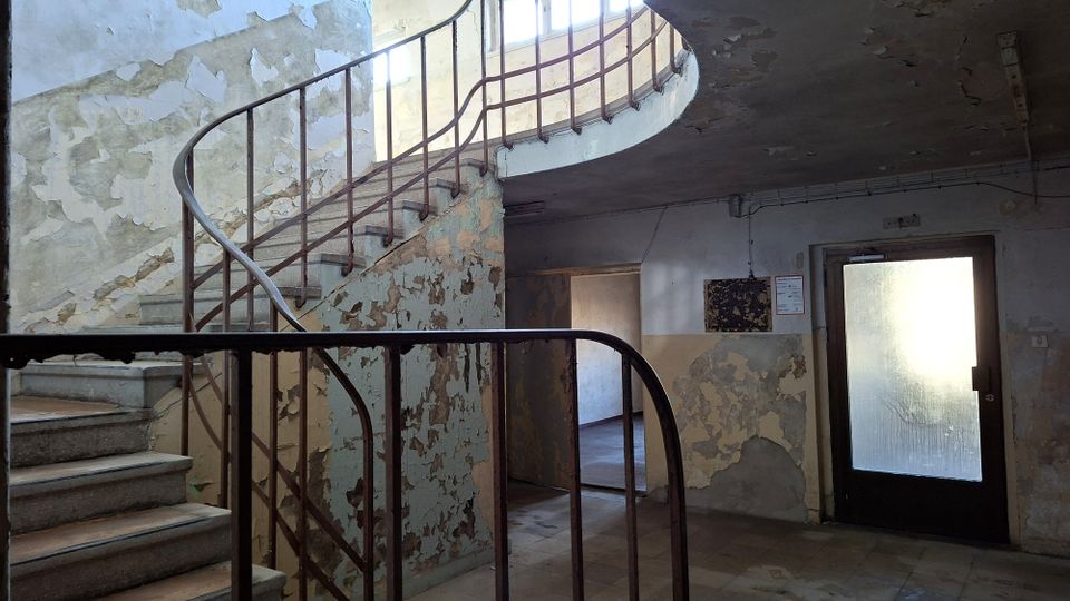 Innenraum eines heruntergekommenen Treppenhauses mit geschwungener Steintreppe und rostigem Metallgeländer, die zu einer oberen Galerie führt. Abgeplatzter Putz an den Wänden und eine Tür mit Milchglasfenster prägen die karge, ältere Gebäudesituation.