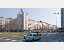 Weitläufige Straße mit historischem Wohngebäude im Stil des Sozialistischen Klassizismus; das Gebäude ist mehrstöckig, mit Balkonen, großen Fenstern und einem Werbegerüst auf dem Dach. Im Vordergrund fahren Autos, darunter ein blauer Trabant, und eine Grünfläche mit kahlen Bäumen grenzt an die Straße.