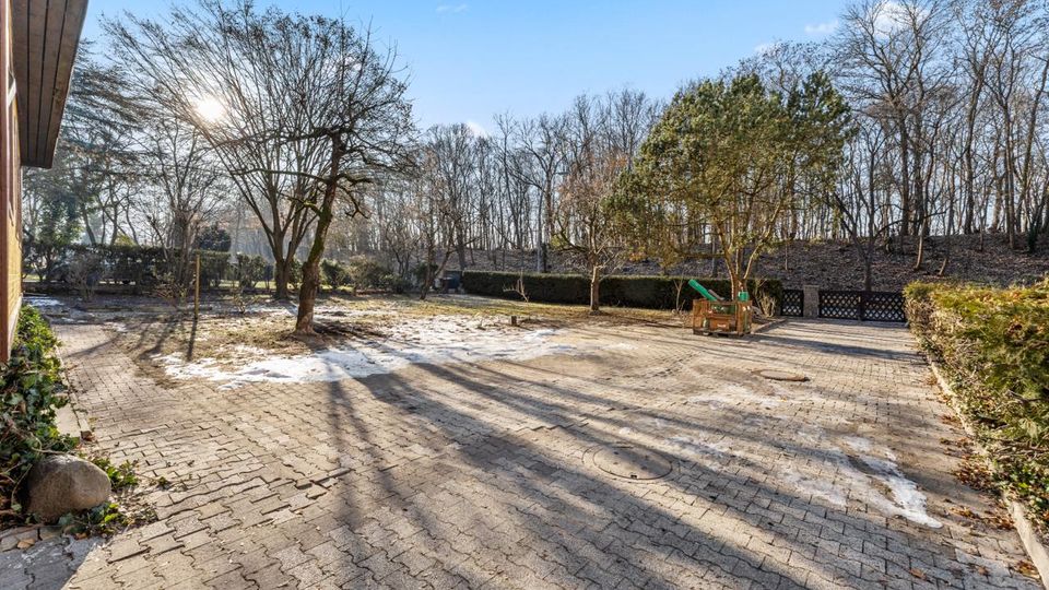 Großzügiger, gepflasterter Hof mit vereinzelten Schneeresten und langen Schatten, umgeben von Hecken und kahlen Bäumen. Im Hintergrund liegt ein bewaldeter Hang, rechts steht eine kleine Holzkonstruktion mit Rutsche.