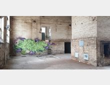 Verlassener Industrie-Innenraum mit freiliegenden Backsteinwänden, einem großen gemauerten Kamin und einer Wand mit buntem Graffiti. Der Raum ist spärlich beleuchtet, zeigt teilweise beschädigte Böden und Fenster sowie offene Durchgänge.