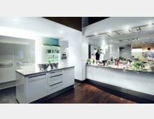 Moderne, professionelle Küche mit weißen Hochglanzschränken, mehreren Edelstahl-Arbeitsflächen und integrierten Geräten. Im Vordergrund stehen große Schüsseln und Gläser auf einer Kücheninsel, im Hintergrund sind offene Regale mit Weinflaschen und Kräutern sowie Personen beim Zubereiten von Speisen zu sehen.