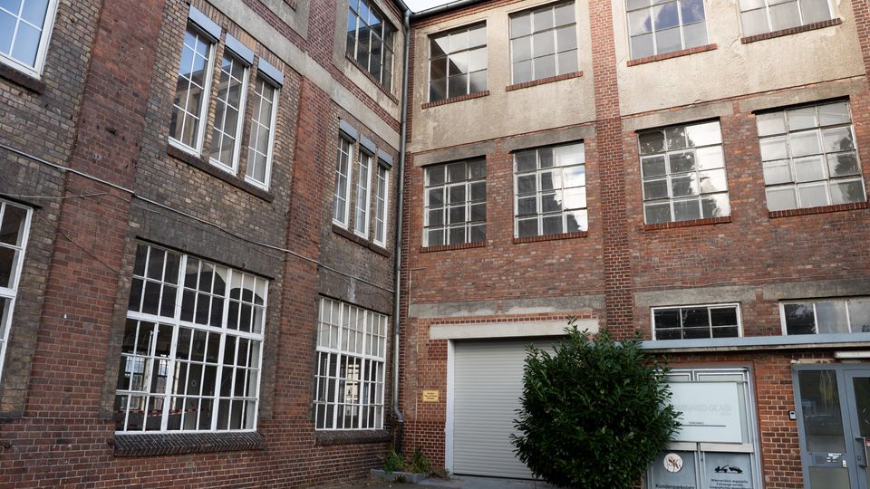 Innenhof eines älteren Industrie- oder Fabrikgebäudes aus rotem Backstein mit großen, vielfach unterteilten Fensterfronten und verputzten Wandbereichen. Unten sind ein Rolltor, eine Seitentür und ein kleiner Strauch vor der Fassade zu sehen.