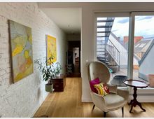 Wohnzimmer mit heller Ziegelwand, an der zwei abstrakte Gemälde hängen. Im Vordergrund ein beigefarbener Sessel mit bunten Kissen, ein kleiner Holztisch und eine große Fensterfront, dahinter eine Dachterrasse mit Außentreppe und Pflanzkübeln.