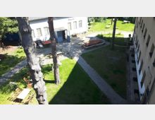 Hofansicht mit Rasenflächen, gepflasterten Wegen und vereinzelten Bäumen. Umgeben von einem weißen Gebäude mit Fenstern und einem beigen Gebäude auf der rechten Seite; mehrere Sitzmöglichkeiten und Holztische sind im Hof aufgestellt.