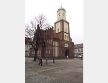 Historisches Backsteingebäude mit gotischen Elementen und Kirchturm, das eine offene Durchgangspforte zeigt; davor ein gepflasterter Platz mit vereinzelten Fußgängern, Laternen und umliegenden Altbauten.