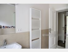 Modernes Badezimmer mit hellbeigen Fliesen an der unteren Wand, einem weißen Waschbecken, einem großen Wandspiegel, einem weißen Handtuchheizkörper und einer offenen weißen Tür, die in einen benachbarten Raum führt. Im Spiegel sind verschiedene Pflegeprodukte sichtbar.