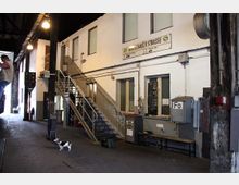 Innenansicht eines Bahnhofsgebäudes mit einer Metalltreppe, die zu einer oberen Ebene führt. An der Wand befinden sich vergitterte Fenster, ein Schild mit der Aufschrift „Warschauer Straße“ sowie verschiedene technische Kästen und Schalter; im Vordergrund steht ein kleiner Hund.