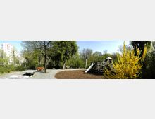 Ein Spielplatz mit Holz-Klettergerüst, Rutsche und umgebender Mulchfläche, umgeben von Bäumen und gelben Blütensträuchern. Im Hintergrund sind ein mehrstöckiges Gebäude und eine Tischtennisplatte zu sehen.