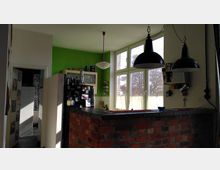 Wohnküche mit einer Theke aus rotem Backstein, darüber hängen zwei schwarze Hängelampen. Im Hintergrund ein grüner Wandabschnitt, ein Fenster mit Blick ins Freie und eine Küche mit schwarzem Kühlschrank sowie Regalen. Links führt eine Tür in einen angrenzenden Raum mit Fliesen.