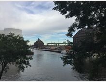 Ein Fluss in einer Stadtlandschaft, umgeben von Industriebauten und alten Backsteingebäuden. Im Vordergrund stehen Bäume, während der Hintergrund moderne Brückenstrukturen und ein altes Speichergebäude zeigt.