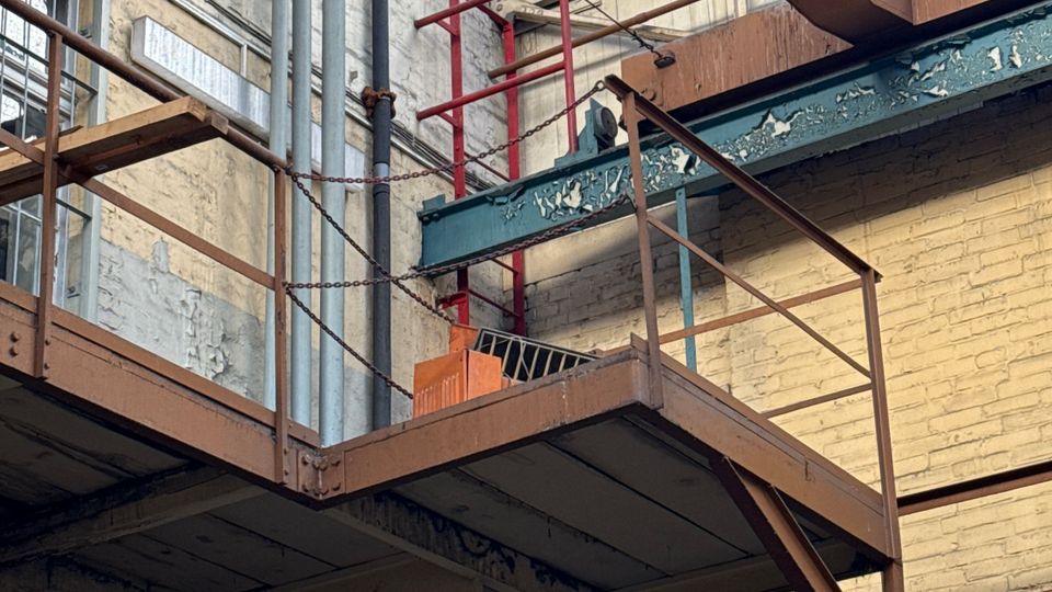 Blick in einen industriellen Innenhof mit Stahlstegen und Treppen, freiliegenden Rohrleitungen und Backstein- sowie Betonwänden. Oben überspannt ein Metallrahmen mit Drahtgitter das offene Dach und lässt den Himmel sichtbar.