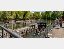 Kanu in einer Schleuse auf einem Fluss, umgeben von grünen Bäumen und einem Uferbereich mit Restaurants und Sitzbereichen unter Sonnenschirmen. Mehrere Personen stehen oder sitzen am Ufer und beobachten die Szene.