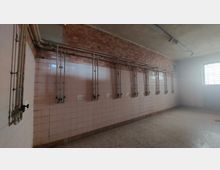 Ein leerer Raum mit kahlen Wänden aus rosa Fliesen und sichtbaren Wasserleitungen, die vertikal entlang der Wände verlaufen. Die Decke ist aus Beton, und ein vergittertes Fenster auf der rechten Seite lässt Tageslicht herein.