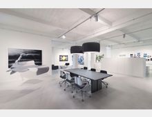 Moderner, minimalistischer Büro- und Besprechungsraum mit einem großen Tisch und acht Drehstühlen, weißen Wänden, abstrakten Kunstwerken an den Wänden und einer auffälligen Hängeskulptur im Vordergrund. Die Beleuchtung erfolgt durch schwarze, zylindrische Pendelleuchten, und das Ambiente wirkt hell durch die offene Gestaltung und das helle Material.