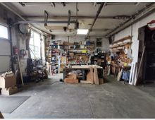 Eine Werkstatt mit Betonboden und weiß gestrichenen Ziegelwänden. Im Raum befinden sich ein zentraler Arbeitstisch, Regale mit Werkzeugen und Materialien an den Wänden sowie Holzplatten und Maschinen. Ein großes Fenster links sorgt für Tageslicht.