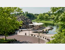 Eine Außenansicht eines Cafés mit einer gewölbten, begrünten Dachkonstruktion neben einem Teich. Rund um das Café befinden sich Sitzbereiche mit Sonnenschirmen, Spazierwege und grüne Bäume.